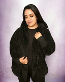 A female model wearing a black teddy hooded sherpa jacket with a relaxed collar neckline, standing against a purple background.